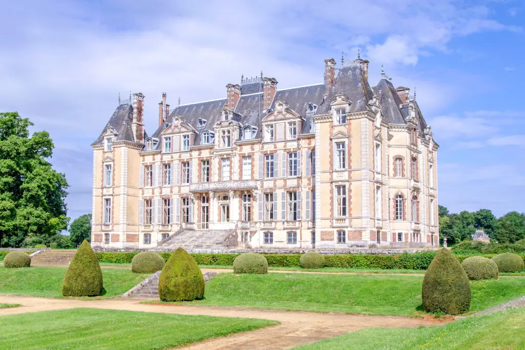 A grand, historic château with a slate roof and ornate stone facade stands amidst manicured lawns near Le Mans. This elegant estate, perfect for an exclusive accommodation during the 24H Le Mans, features tall windows and intricate architectural details against a clear blue sky.