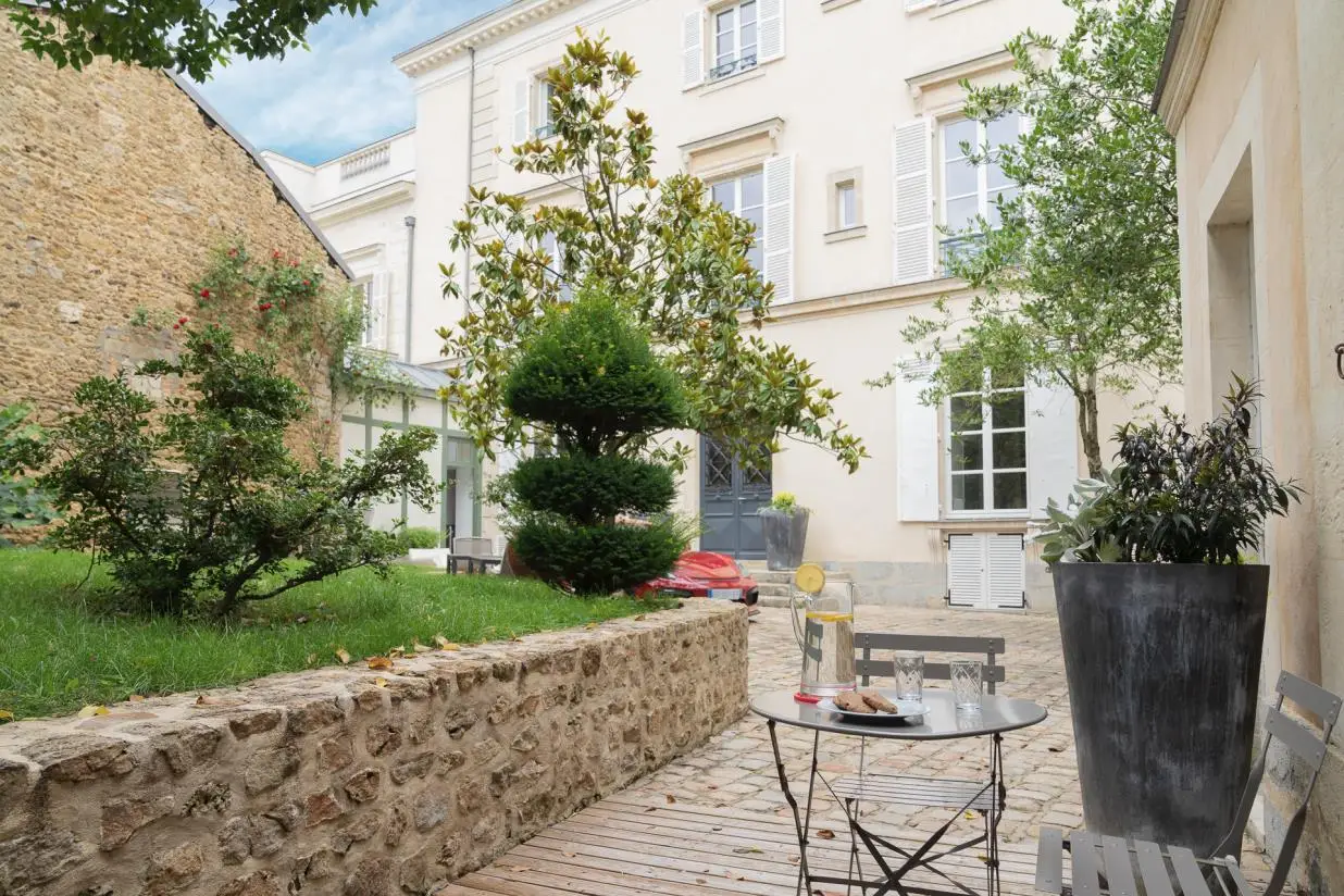 A cozy courtyard at a luxury French manor near the 24H Le Mans, with brick and stone walls, potted plants, trees, a grassy patch, and a wooden deck. Enjoy exclusive hospitality with metal table and chairs by the window under partly cloudy skies.