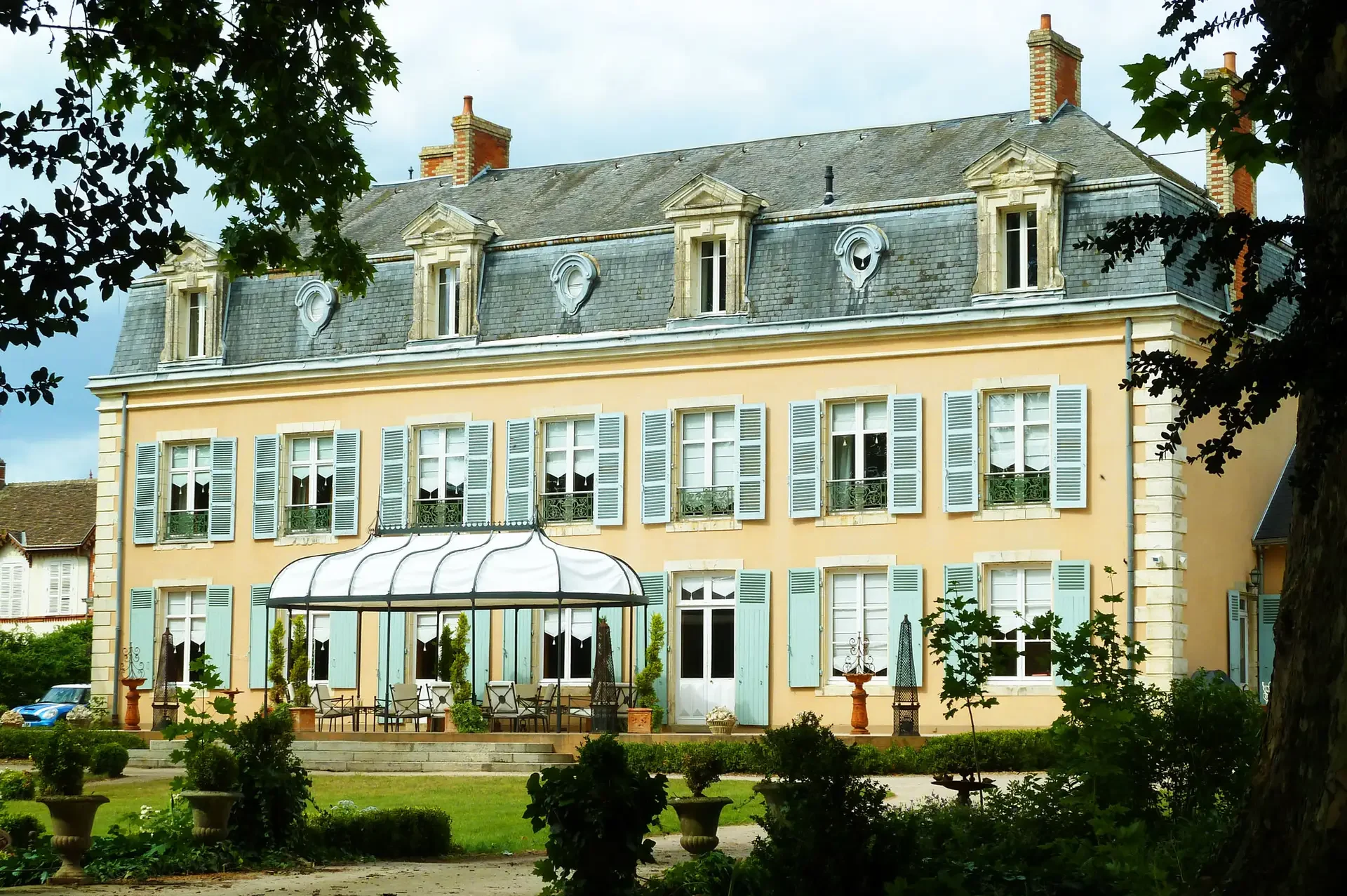 A large, elegant château near Le Mans, boasting yellow walls and blue shutters, features a glass conservatory at its center. Surrounded by manicured gardens and trees under an overcast sky, it offers a serene and luxurious stay near the 24H Le Mans race.