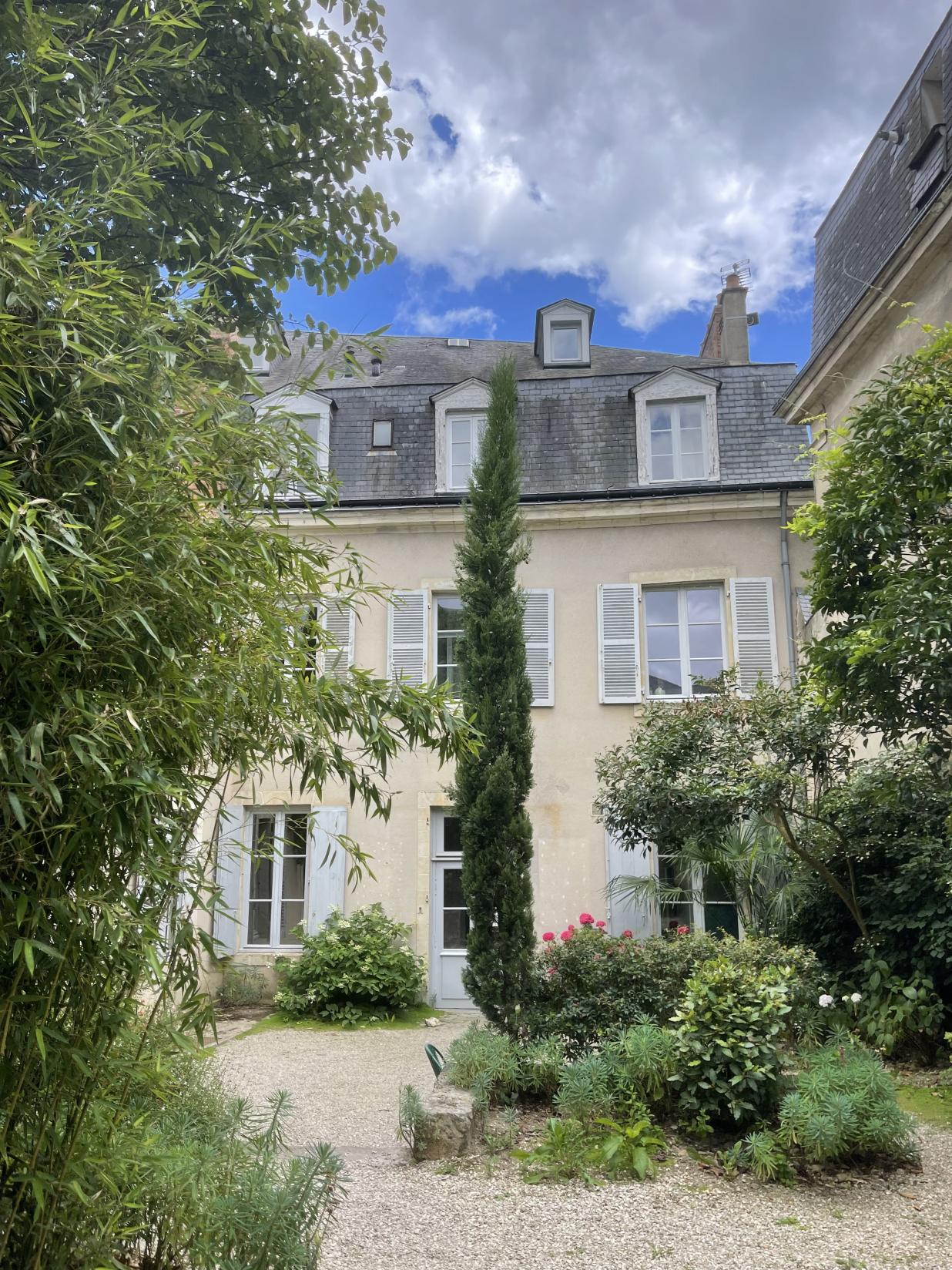 La Villa des Arts, a cream-colored building with gray shutters and a slate roof, stands behind a lush garden filled with green plants, a tall cypress tree, and a gravel path under a partly cloudy sky.